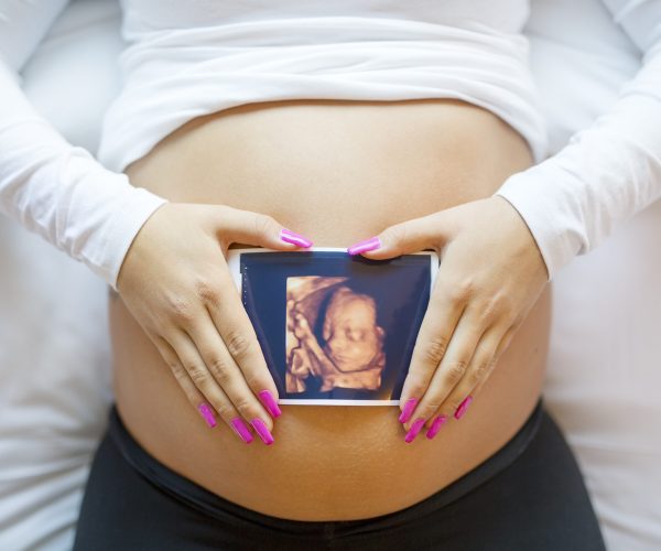 Close-up of an pregnant woman holding ultrasound photograph of a baby in front of the swollen bare tummy. Laying in the bed.